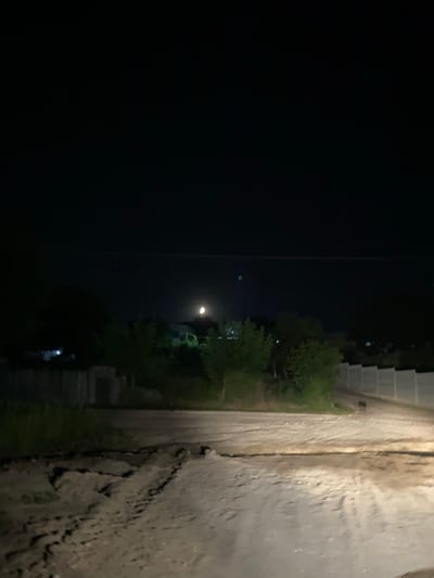 The image captures a nighttime scene in Negrești, Moldova. The perspective is from a ground-level view, looking across a narrow, rough dirt path. To the right of the path, a textured, uneven embankment rises, appearing to be made of packed earth or gravel with visible erosion and debris. The path itself is a lighter color, suggesting it's made of dirt or fine gravel.
To the left of the path, a dark, open expanse dominates the left side of the frame, suggesting a void or very dark area, possibly a field or undeveloped land. In the middle distance, beyond the path, several trees with dense foliage are visible, illuminated by artificial lights. These lights create a faint glow in the darkness. Further in the distance, faint outlines of buildings or structures can be discerned, some with hints of lights. One particular light source in the distance appears to be a streetlamp or a bright overhead light, casting a visible beam into the dark sky. The overall atmosphere is dark and quiet, typical of a rural nighttime setting. There is no visible text in the image. The time of day is clearly night, and no specific weather conditions are apparent.