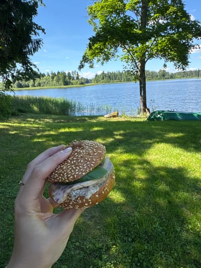 A person's hand, adorned with a ring on the ring finger, holds a sandwich consisting of a sesame seed bun with filling. The sandwich is held in the foreground against a backdrop of a body of water, likely a lake, in Lendži, Latvia. The water is calm and reflects the blue sky, with green reeds lining the shore. Beyond the water, a dense forest of trees is visible. In the middle ground, a tall tree stands to the right, and a green canoe rests on the grassy bank. The foreground is a well-maintained lawn with shadows cast by the surrounding trees, suggesting a sunny day. The overall scene depicts a serene outdoor setting ideal for a picnic.