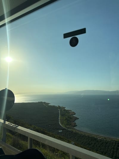 This image captures a expansive coastal landscape under a bright, clear sky, likely viewed from an elevated position such as a window, bridge, or high platform. The scene is dominated by a large body of water, presumably Iznik Lake given the provided context, which stretches across the right and central parts of the frame. The water's surface reflects the bright sunlight, creating shimmering patterns.

Along the water's edge, a verdant coastline unfolds, characterized by dense green vegetation and rugged terrain, including some rocky formations. A narrow, winding road is visible, tracing the contours of the shoreline and disappearing into the distance. In the background, hazy mountains or hills line the horizon, adding depth to the landscape.

The sun, a brilliant white orb with noticeable lens flare, is low in the sky towards the lower left of the image, indicating either sunrise or sunset. The sky transitions from a light blue at the top to a warmer, yellowish hue near the horizon, illuminated by the low-hanging sun.

In the foreground, on the right side, a substantial gray structural element, possibly a beam or railing, extends diagonally across the frame, suggesting the viewpoint is from a man-made structure. On what appears to be a glass surface (perhaps a window), a dark, rectangular object and a smaller, dark circular object with an indistinct symbol are visible in the upper left. Another dark circular object, possibly part of a vehicle's interior or mirror, is partially visible in the lower right.

The image conveys a sense of tranquility and natural beauty, showcasing a serene moment in a scenic location. No people are visible, and no discernible text can be read.