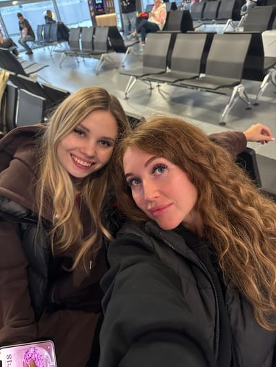 Two young women are posing for a selfie. One woman has long, blonde hair and is wearing a brown jacket. The other woman has long, curly brown hair and is wearing a black jacket. They are both smiling. The background appears to be an airport waiting area, with rows of seats and people in the distance. The location in the city of Boldino, Russia is not visually apparent in this image. A phone with a purple screen is visible in the bottom right corner of the image.