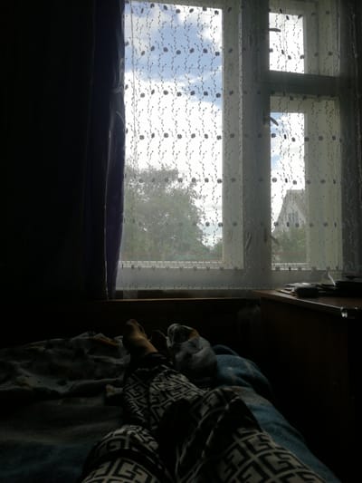 The image captures a view from a person's perspective, lying down on a bed. The subject is focused on their legs, which are covered in black and white patterned pajama bottoms, resting on a blue and gray patterned blanket. The person's feet are also visible.
To the right of the bed, a wooden desk or dresser is partially visible, with a few books or papers stacked on it. A dark curtain hangs on the left side of the frame, obscuring much of the room.
The main focus beyond the immediate foreground is a window, partially covered by a sheer, white lace curtain with a delicate floral pattern. Through the curtain, a bright, partly cloudy sky can be seen. Outside the window, trees are visible, suggesting a rural or suburban setting. In the distance, a house with a pitched roof is discernible.
The lighting suggests it is daytime, likely morning or afternoon, given the bright sky. The overall mood appears relaxed and perhaps introspective, with the subject observing the scene outside from a comfortable indoor space. There is no visible text in the image. The location context of Dubrovka, Russia, implies a setting in a Russian village or town.