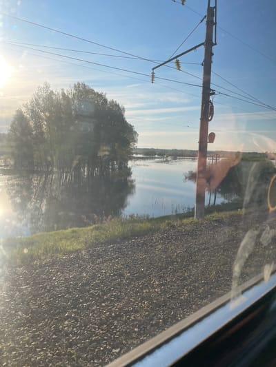 The image shows a view from inside a vehicle, likely a car, in Talmenka, Russia. The roadside is visible with grass and gravel. An electrical pole stands with wires running along it. Reflections of trees and sky are prominent on the glass, obscuring the background details. The sky is blue with streaky clouds.