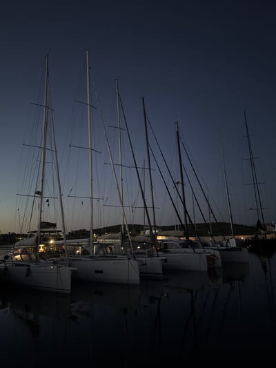 This image presents a forensic depiction of a marina at twilight or early night in Lavreotiki Municipal Unit, Greece. A row of approximately eight sailing vessels, predominantly monohulls but including at least two catamarans, are moored side-by-side. Their tall, slender masts and intricate rigging create a dense pattern against the deep indigo-blue sky, which exhibits a subtle gradient from lighter blue-grey near the horizon to darker overhead, with a few faint star-like points visible in the upper right.

The boats feature white hulls, with details such as "BV25500" visible on one monohull and "4VE9300" on a catamaran. One catamaran also displays a distinct orange logo or graphic on its transom. Warm yellow light emanates from the interior cabins of several vessels, illuminating their structures. The water in the foreground is exceptionally calm and dark, acting as a mirror that provides clear, inverted reflections of the boats, their masts, and the artificial lights. In the background, low-lying hills or land masses are visible, outlined against the sky, with scattered artificial lights indicating distant structures or habitation. No people are visible within the scene.