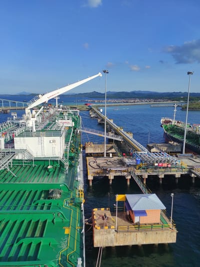 This media file presents a bright, clear-day aerial view of an industrial port in Kunak, Malaysia, depicting a large green tanker ship docked at a concrete pier. The scene is set against a backdrop of deep blue water and a distant coastline featuring mountains and scattered urban development under a partly cloudy sky.

In the foreground, the extensive deck of the green tanker ship is visible, adorned with numerous white structures, intricate piping, and various pieces of machinery. A prominent white crane boom extends from the ship, displaying the text "INGERSOLL SWR 15T@12~25M," indicating its specifications. On the ship's deck, "NO SMOKING" and "SAFETY FIRST" signs are clearly visible, alongside yellow painted lines with text like "FLAP ACCESS POINT KEEP CLEAR," "SAFE AREA," and "DANGEROUS AREA."

The ship is securely moored to a concrete pier, which supports a complex system of yellow, blue, and grey pipelines, access walkways, and stairs. These pipelines connect to the ship, suggesting ongoing or preparatory liquid transfer operations, likely for loading or unloading petroleum products or chemicals. Further down the pier, another, smaller dark-hulled ship is also docked. A small, multi-colored building with a blue roof and orange and blue walls is situated on a section of the pier. Several tall poles with floodlights are scattered throughout the facility, implying capability for continuous operations. The overall impression is of a bustling industrial maritime facility under sunny weather conditions.