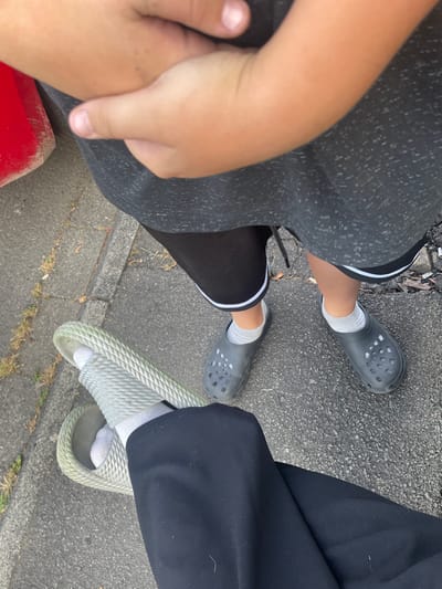 The image depicts two individuals' legs and feet on an asphalt surface, possibly a sidewalk, adjacent to a curb with some sparse vegetation. The person on the right wears black Croc-style shoes with grey socks and dark shorts with white trim. The person on the left wears a light-colored woven sandal over grey socks, and black pants. A portion of a red object, possibly a bucket or container, is visible in the upper left corner. The overall scene suggests an outdoor urban or suburban environment. The location in the city of Wiehl, Germany is not visually discernible from the provided image.