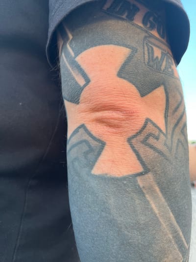 The image shows a close-up of a person's elbow with a large tattoo. The tattoo is predominantly black and grey, with a light-colored, cross-like design centered on the elbow. The skin on the elbow itself appears slightly reddened and has some texture. A portion of a dark t-shirt is visible on the left side of the image. The background is out of focus, but suggests an outdoor setting with a light blue sky. There is no discernible information about the city of Palanca, Moldova, within the image itself.