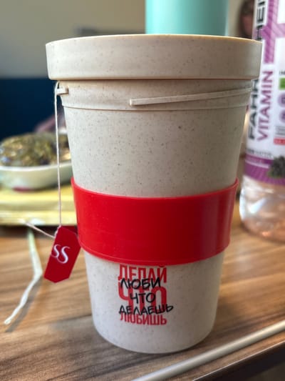 A beige, speckled reusable cup with a matching lid is centrally positioned on a light-brown wooden table. A red silicone band encircles the cup's middle. Below the band, Russian text is printed: "ДЕЛАЙ ЛЮБИ ЧТО ДЕЛАЕШЬ ЛЮБИШЬ," which translates to "DO WHAT YOU LOVE LOVE WHAT YOU DO." A white string extends from a slot in the lid, connected to a small red tag featuring white "SS" lettering, indicating a tea bag. In the blurred background to the left, a white bowl containing dark, textured contents is visible. To the right, a clear plastic bottle with a purple label partially displays "FAIRTRADE VITAMIN WATER" and an image of dark berries. A faint, indistinct reflection of a human figure is discernible behind the bottle. A thin, light-colored rod rests on the table surface in the lower right foreground. This scene is located in Krasnoufimsk, Russia.