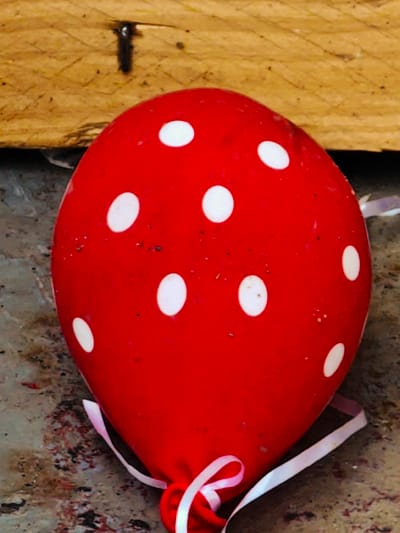 A red balloon with white polka dots is resting on a speckled gray surface, positioned in front of a rough wooden plank with a nail hammered into it. A thin white ribbon is tied around the neck of the balloon and trails to the right. The scene is outdoors, with the background suggesting an urban or industrial setting. This location is Erkalovo, Russia.