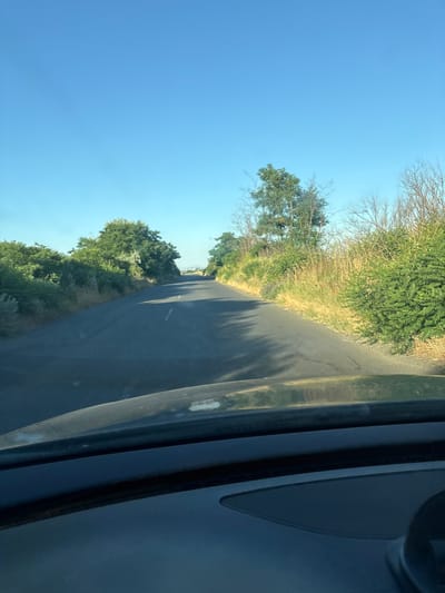 This is a first-person perspective shot taken from inside a vehicle, showing a road leading into the distance. The road is paved and has a white dashed line marking the center. On either side of the road are grassy verges with dense green foliage and trees. The sky is clear and bright blue. The image appears to have been taken during the daytime, possibly in the late afternoon, given the angle of the shadows cast on the road. The overall environment suggests a rural or semi-rural setting. There are no people or specific landmarks visible that would definitively place the location in Kableshkovo, Bulgaria.