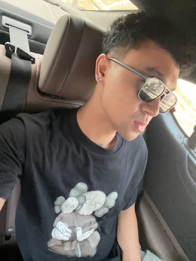 The image captures a scene inside a vehicle, located within the city of Kayuambon, Indonesia. A man is depicted sitting in what appears to be the back seat, looking downwards and slightly to his right. He has short, dark, curly hair with faded sides. He is wearing reflective, rectangular-framed sunglasses that obscure his eyes. A small, circular stud earring is visible in his left ear. His lips are slightly parted. He wears a black short-sleeved t-shirt featuring a large graphic of two stylized grey and beige figures, characteristic of KAWS artwork, with X's for eyes. Behind his left shoulder, a brown textured car seat headrest and a black seatbelt with its securing mechanism are visible. Bright natural light illuminates the scene from a window to his right, reflecting in his sunglasses. The vehicle's interior trim appears dark-colored.