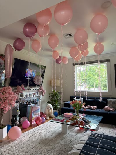 The image depicts a living room decorated for a birthday party. Pink and rose-gold balloons are scattered throughout the room, some attached to the ceiling with ribbons and others resting on the floor. A large television screen displays a birthday graphic with candles. A fireplace mantle is adorned with a "Happy Birthday" banner and various figurines. In the foreground, a plush toy dog sits beside a rainbow-striped gift bag. A coffee table in the center of the room holds a vase of tulips, a toy, and gift boxes. A dark blue sofa is visible in the background, and a large window offers a view of green trees outside. The room appears to be furnished with a plush rug and a sectional sofa.
