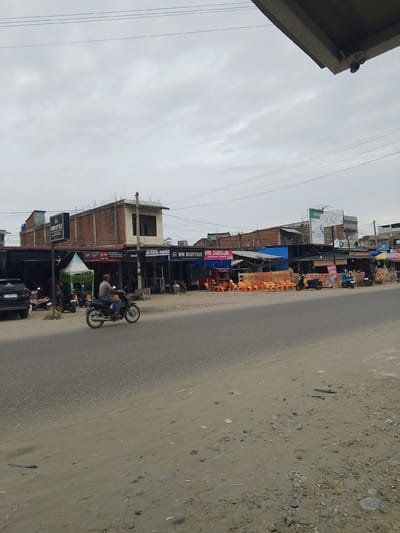 The image depicts a street scene in Gampongpaya, Indonesia, under an overcast sky. A two-lane asphalt road occupies the foreground, with a sandy and somewhat littered shoulder on the right. Along the left side of the road, a row of small commercial establishments with awnings and signage are visible. A black SUV is parked on the left. Several motorcycles are parked in front of the shops, and one motorcycle is in motion, ridden by a man wearing a grey shirt. The buildings appear to be a mix of brick and simpler structures, some with upper floors. Signs for businesses such as "AMRI STYLE," "MAYA MODA," and "WM BOUTIQUE" are discernible. On the right side of the road, several orange plastic chairs are set up outside a food stall, with banners advertising "ES TELUR CREAM." The overall impression is a moderately busy street in a commercial area.