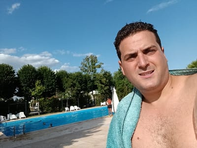A man takes a selfie poolside on a sunny day. He is shirtless with a blue towel draped over his shoulder. Behind him is a blue swimming pool with a few people swimming or standing in it. Lounge chairs line the pool deck. Lush green trees form a backdrop under a blue sky dotted with light, scattered clouds.