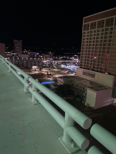The image presents an elevated, downward-angled nighttime view of downtown Reno, Nevada, United States of America.

In the foreground, a white, multi-bar metal railing runs diagonally from the lower left to the center-right, partially obscuring the immediate scene below. The flat, light-colored concrete surface beneath the railing displays a network of fine cracks.

The midground features the Atlantis Casino Resort Spa, a prominent, multi-story building with a reddish-pink facade and a grid of square windows, located on the right side of the frame. A large sign bearing the text "BEST BEST IN THE WEST" is affixed to a lower section of the Atlantis structure. Adjacent and below the Atlantis, a smaller building appears to be under construction or renovation, evidenced by visible scaffolding and white tarping. City streets are visible, characterized by elongated streaks of blue and white light, indicating moving vehicles during a long exposure. Other multi-story buildings, some with illuminated windows, are scattered throughout the midground. Several light sources, including streetlights and blue architectural lighting, illuminate the urban environment.

The background consists of a vast expanse of city lights receding into the distance, signifying a developed urban area. The sky above is dark and featureless. No individuals or discernible specific actions are visible within the frame.