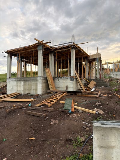 The image shows a building under construction in what appears to be a rural area. The building is a two-story structure with concrete pillars and wooden framing. Rebar extends from the top of the structure. Scaffolding and other construction materials are scattered around the site. There are some other building under construction in the distance. The location is unidentified.