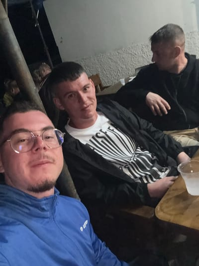 Three men are indoors. One wears a black hoodie, another a black jacket over a white shirt with black lines, and the closest man wears a blue jacket and eyeglasses. The location is potentially Pripek, Bulgaria.