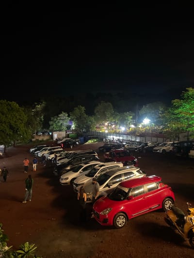 A night-time aerial view shows a busy parking lot in Vagator, India, filled with cars and a few motorcycles. Several individuals are seen walking among the vehicles. In the foreground, a vibrant red hatchback is parked, accompanied by a yellow scooter to its right. The parking lot itself is a dusty, uneven dirt surface. Beyond the immediate parking area, a line of cars, mostly white SUVs and sedans, are neatly parked. In the mid-ground, a row of parked motorcycles suggests a gathering or event. The background is a dense cluster of trees, some illuminated by green lights, and beyond that, the dark silhouette of hills under a night sky. A few structures are visible in the distance, with lights indicating activity. The overall impression is one of a lively outdoor space at night, likely a venue for entertainment or dining, given the parking lot's full occupancy.
