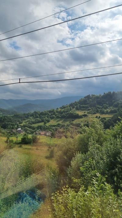 The image depicts a rural landscape in Volosianka, Ukraine. In the foreground, power lines are visible, stretching horizontally across the frame. The middle ground features a rolling green hillside dotted with numerous trees and a small cluster of houses with red and white roofs. A few fields are also visible, some with dry grass. The background consists of a series of undulating, tree-covered mountains under a cloudy sky. The overall impression is one of a serene, natural environment with a hint of human habitation.