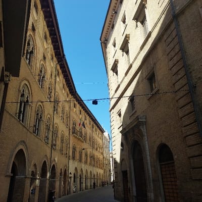 The image displays a view of Via della Loggia in Ancona, Italy, under a clear blue sky with direct sunlight illuminating the scene from the upper left. The street is a narrow, paved thoroughfare, receding into the distance.

On the left, the multi-story Palazzo degli Anziani presents a light-colored brick and stone facade. Its ground floor features a continuous arcade of pointed arches, while upper floors are adorned with numerous windows exhibiting intricate Gothic tracery. Italian, European Union, and a third, indistinct flag are displayed from an upper-story window or balcony. A dark-colored scooter is partially visible parked under the arcade on the left.

On the right, a multi-story building, constructed of similar light-colored brick, features simpler, rectangular windows and two large arched ground-level doorways. The upper portion of this building's facade is brightly lit by sunlight, while its lower portion and right side are cast in shadow. A dark vertical conduit or pipe extends down its right edge.

Overhead, two thin, dark wires or cables stretch horizontally across the street between the buildings. A small, dark rectangular device is suspended from one of these wires. Several indistinct human figures are observed walking away from the viewer in the distant section of the street.