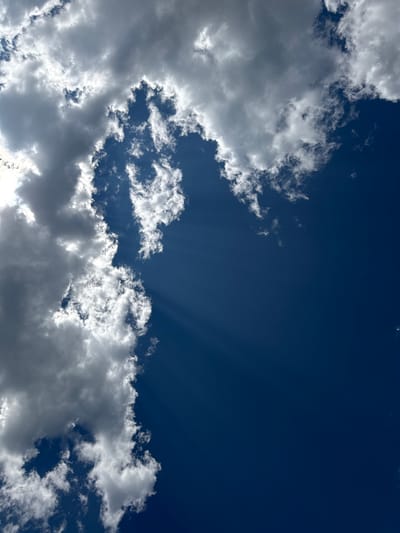 The media file is a photograph of a bright blue sky with scattered white and gray clouds. The clouds are dynamic, with some appearing thin and wispy, while others are thick and volumetric, casting shadows on themselves and the sky. Sunbeams, also known as crepuscular rays, are visible radiating from behind a cloud formation in the upper right quadrant of the image, indicating the presence of sunlight. The overall impression is one of a clear, possibly breezy day with a mix of sunlight and cloud cover. The image appears to be taken outdoors, looking upwards. No people, objects, text, or specific location cues are visible, apart from the implied sky. The time of day is likely daytime, given the brightness of the sky and the sunbeams. The weather is characterized by the presence of clouds, but the intensity of the blue sky suggests it is not overcast.