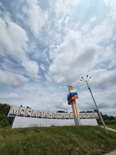 The image shows a sign that reads "МОСКВА-ВЛАДИВОСТОК" in large, orange, three-dimensional letters against a grey background. The sign is placed in a grassy area next to a road, with trees and foliage visible in the background. A lamppost with multiple lights is positioned above and behind the sign. The sky is cloudy with patches of blue. The orientation of the image is rotated, suggesting it was taken from a low angle or the camera was tilted. The text on the sign refers to the cities of Moscow and Vladivostok, which are widely separated locations in Russia. This type of sign is often found at the beginning or end of a long journey or route. The overall scene suggests an outdoor setting on a partly cloudy day, possibly indicating the start or end of a long road trip. No people are visible in the image.