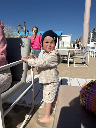 The image, taken on a sunny day with a clear blue sky, shows a young child in a beachside or terrace setting, likely in Middelkerke, Belgium. In the foreground, a toddler, wearing a dark blue beanie with a white "B" logo and a light-colored linen-like shirt and shorts, is standing. The child is smiling faintly, holding onto the white frame of a grey outdoor armchair. Their bare feet are visible on the sandy ground, suggesting they might be just off a sandy beach or on a raised deck with sand nearby.
Partially visible behind the child to the left is a person with blonde hair tied in a ponytail, wearing a bright pink top. Further in the background, a series of white buildings, possibly hotels or apartments, line the coast. White outdoor dining furniture (tables and chairs) are scattered across what appears to be a sandy or wooden decking area. A large, cream-colored sun umbrella pole is also visible. On the far right, a small section of a patterned, possibly crocheted, item (perhaps a bag or hat) sits on an upholstered chair. Shadows are cast by the furniture, indicating bright sunlight, likely mid-day to late afternoon. The overall scene conveys a relaxed, pleasant atmosphere at a coastal location.