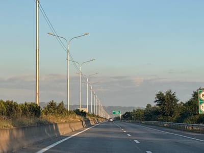 This is a medium-distance, eye-level, horizontal shot of a highway in Dzveli Kanda, Georgia. The highway has multiple lanes, with a white line marking the lanes. On the left side of the highway, there is a concrete barrier followed by a row of trees and then tall streetlights. On the right side of the highway, there are speed limit signs with the numbers 80 and 110 in red circles within green signs. The sky is clear and blue, with some clouds visible in the distance. There are a few cars on the highway. The overall impression is one of a modern highway in a rural or semi-rural setting.