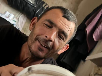 A medium shot captures a man from the chest up, looking directly at the camera with a slight smile. He has short, dark hair, a receding hairline, and visible wrinkles on his forehead and around his eyes. He sports a mustache and stubble. His left ear is visible, and his right ear is mostly obscured. He is wearing a dark t-shirt. In the background, to the left, a black bag with a white stripe is visible. To the right, a purple garment is hanging. The environment appears to be indoors, possibly a residential setting, with textured walls and a light-colored surface in the foreground that could be a table or a similar piece of furniture. The lighting suggests an indoor environment.