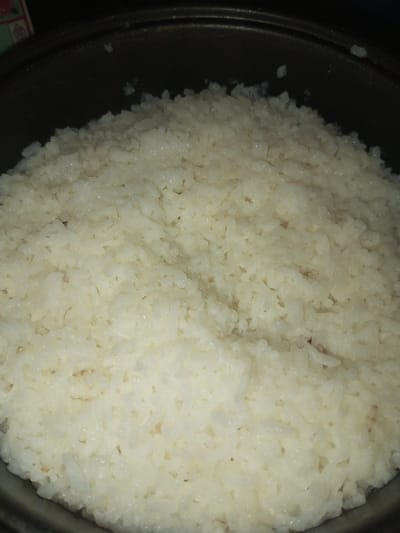 The image displays a quantity of cooked white rice contained within a dark-colored pot, observed from a slightly elevated, close-up perspective. The rice occupies the majority of the frame, appearing moist with visible individual grains, exhibiting a uniform off-white to pale yellow coloration. Sparse, small, dark flecks are distributed throughout the rice mass. The pot's dark, possibly non-stick, rim is visible along the top and sides of the rice. No individuals, actions, or specific environmental details beyond the immediate container are discernible within the frame. The scene is located in Doramamidi, India.