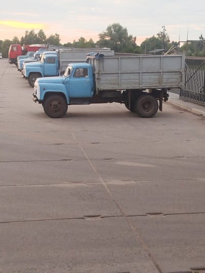 In Krolevets, Ukraine, a line of parked blue flatbed trucks with grey cargo beds are visible on a paved lot. The trucks are parked in a row, facing to the right, with their beds covered by a grey tarp. The foreground shows the pavement with visible cracks and seams. In the background, trees and a fence are visible. The sky is overcast with hints of sunset colors. No people are present in the image.