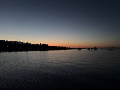 This image captures a serene twilight scene at the shore of a large body of water, likely a lake, given the context of Sary-Oy, Kyrgyzstan. The sky transitions from a deep, dusky blue at the top to a soft gradient of pale orange and yellow near the horizon, indicating either sunrise or sunset. A dark, silhouetted treeline stretches across the distant shore, interspersed with what appear to be scattered lights, suggesting a settlement or resort area. In the middle ground, several piers with structures on them extend into the water, appearing as dark shapes against the glowing horizon. The water itself is calm with subtle ripples, reflecting the dim light and the dark forms of the piers and the shoreline. The overall mood is peaceful and tranquil, evoking the quiet beauty of dusk by the water. There are no visible people or text in the image.