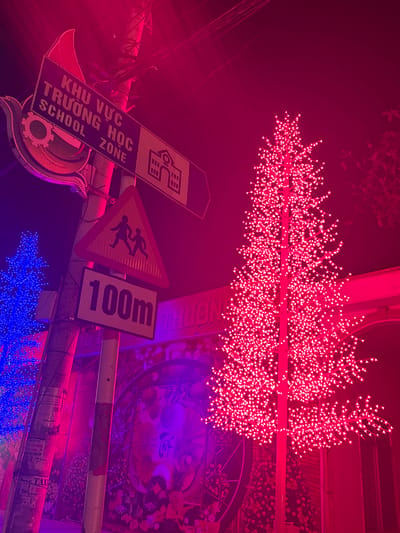 This nighttime scene is located in Quỳnh Lưu Commune, Vietnam, featuring a street illuminated predominantly by red and blue artificial light. In the foreground, a metal utility pole supports several signs. A horizontal black sign reads "KHU VỰC TRƯỜNG HỌC SCHOOL ZONE" in white text, with an arrow pointing right towards a white school building icon. Below this, a yellow triangular warning sign displays two black silhouettes of children walking. Further down, a white rectangular sign indicates "100m" in black text.
To the right of the pole, a tall, slender, bare tree-like structure is entirely covered in numerous small, intensely red lights. A similar, partially visible tree-like structure, illuminated with blue lights, is present to the left of the pole. The background features a large architectural element, possibly an archway or building facade, adorned with colorful murals or posters and bathed in purple and pink light. Overhead electrical wires are discernible at the top of the frame. The sky is dark, indicating night.