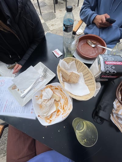 A high-angle shot reveals a table covered with remnants of a meal. A person in a blue hoodie holds a phone on the left side of the table, near a brown bowl and a water bottle. A "CATUNAMBU El café de Andalucía" container sits next to a wicker basket holding pieces of bread and white napkins. A plate smeared with sauce and food scraps, also containing crumpled napkins, sits next to a square dish with cutlery and a menu listing food items and prices. A near-empty glass and a handbag are situated towards the right of the table. A person in a black jacket is visible on the lower-left side. The setting appears to be a restaurant in Jerez De La Frontera, Spain, based on the menu.
