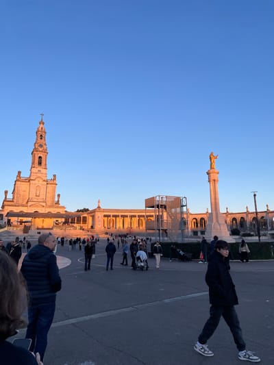 A vertical shot depicts a clear blue sky and the Sanctuary of Fátima at golden hour. The basilica, with its tall spire, and the colonnade building are prominently displayed. People are scattered on the paved area in front. A golden statue stands on a pillar. A person in a black coat and sneakers walks by. The image seems to be taken at ground level, looking up at the buildings and the sky.