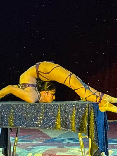 A female acrobat performs on stage at an event located in Tejalpa, Mexico. The acrobat, wearing a gold-colored outfit with purple trim, has her feet pressed against a decorated tower. The acrobat is in a contorted position, with her back arched and head touching the tower. The backdrop is a dark color, with small, irregularly spaced white spots.