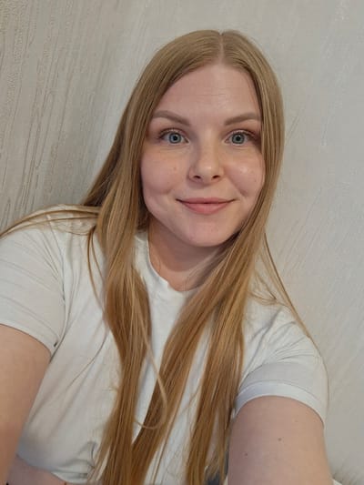 A close-up selfie depicts a young woman with fair skin, freckles, and blonde hair, partially covering a white t-shirt. She has blue eyes and a slight smile. The background appears to be a light-colored wall. There is no evidence to suggest that this picture was taken in the city of Ozyorny, Russia.