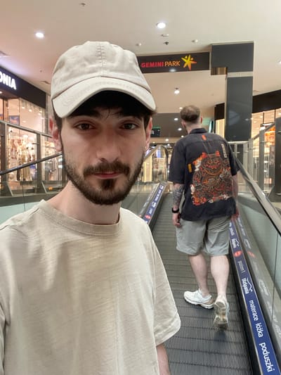 The image shows two people on a shopping mall escalator. The person in the foreground is taking a selfie, wearing a beige baseball cap and a light-colored t-shirt. They have dark hair and a beard. In the background, another person is seen from the back, wearing a black shirt with a colorful, elaborate Japanese-inspired design on it and light gray shorts. They are also wearing white sneakers. The escalator has blue advertising strips with text in Polish. One strip reads "DlaSpania materace łóżka poduszki", which translates to "For Sleeping mattresses beds pillows". Another strip says "poziom 1 Dlaspania" meaning "Level 1 Dlaspania". A vertical sign for "GEMINI PARK" with a yellow and red star logo is visible on the left side of the image. The setting appears to be an indoor shopping mall, possibly during the daytime given the lighting. The overall mood is casual and everyday.