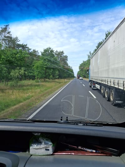 The image depicts a view from inside a vehicle, looking out through the windshield. The scene is a road in Cierpice, Poland, likely during the daytime given the bright sky and clear visibility. The road appears to be a two-lane highway surrounded by lush green trees and vegetation on either side. In the foreground, the dashboard of the vehicle is visible, with a package of tissues and a pen lying on it. The windshield itself has some faint water marks and a clear outline of what appears to be a de-icing or defogging pattern.

On the road, the primary subject is a large, white semi-trailer truck with a blue cab, traveling in the same direction as the viewer's vehicle. The truck has multiple axles and the brand name "KRONE" is visible on its rear. Further down the road, other vehicles, including a smaller truck and a white car, are also visible, suggesting moderate traffic. The sky above is a vibrant blue with scattered white clouds. The overall impression is a snapshot of travel on a rural highway in Poland.