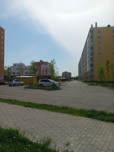 The image depicts an outdoor urban scene in Kommunar, Russia. A paved area stretches into the distance, flanked by green grassy strips and rows of trees. A sidewalk made of gray paving stones is visible in the foreground.
On the left, a multi-story apartment building of orange brick construction is partially visible. Several cars are parked near the base of this building. Further ahead, between the trees, stands a white and red multi-story building.
To the right is a long, yellow apartment building, punctuated by rows of windows. A few trees are planted along its base.
In the distance, a few cars are visible on the road, and another multi-story building can be seen. The sky is pale blue with some cloud cover, suggesting a sunny day. There are no people visible in the image.