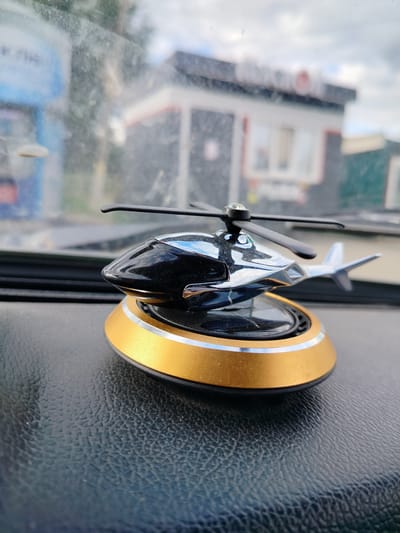 An external, eye-level, close-up shot shows a solar-powered helicopter car air freshener with a gold base resting on a dark, textured dashboard. The helicopter has a shiny black and chrome body and black rotors. The background is blurred, showing a building with windows and a sign, possibly a storefront, under an overcast sky. The image was taken in Grabovo, Russia.