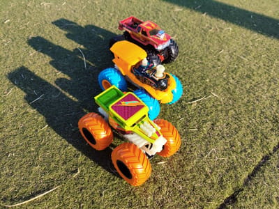 Three toy monster trucks are arranged in a stacked formation on a patch of green, short-cut grass under bright sunlight. The grass appears artificial or very meticulously maintained. Long shadows are cast to the left of the trucks, indicating the sun is positioned to the right and slightly behind the viewer.
The truck at the bottom is predominantly lime green with bright orange, oversized wheels. It features a white chassis and what appears to be a yellow and purple checkered pattern on its hood.
Stacked on top of the green truck is a bright yellow toy truck with large blue wheels. This truck has some black detailing with colorful graphics and white elements that resemble exhaust pipes or chimneys.
The topmost truck is red and yellow, also with large black wheels. It has a visible engine component on the hood and what looks like a chrome accent on the front.
The overall scene is static, with no signs of human activity or interaction with the toys. The setting is an outdoor environment, likely a park or garden, given the grassy surface. The location is specified as Melling, United Kingdom.