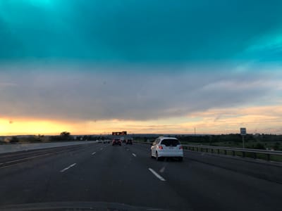 The image captures a view from a vehicle traveling on a highway, likely in King, Canada. The scene depicts a sunset with a dramatic sky transitioning from a bright yellow and orange horizon to a deep turquoise and grey overhead. Wispy clouds are scattered throughout, some reflecting the warm hues of the setting sun.

The highway has multiple lanes, and several vehicles are visible in front, including a white hatchback, a black sedan, and other cars further in the distance. The road is dark asphalt, with white lane markings. A concrete barrier separates the lanes from the opposing traffic. On the right side of the highway, a metal guardrail runs alongside a grassy embankment dotted with trees and distant rolling hills.

A speed limit sign indicating "100" is visible on the right side of the road. Overhead, electronic signs are present, with one showing "WATCH YOUR SPEED" and another displaying a warning symbol and text that is partially obscured but appears to relate to speed limits or surveillance. The overall impression is one of a journey during dusk, with the dynamic sky creating a sense of serenity and natural beauty. The setting suggests a rural or suburban highway environment.