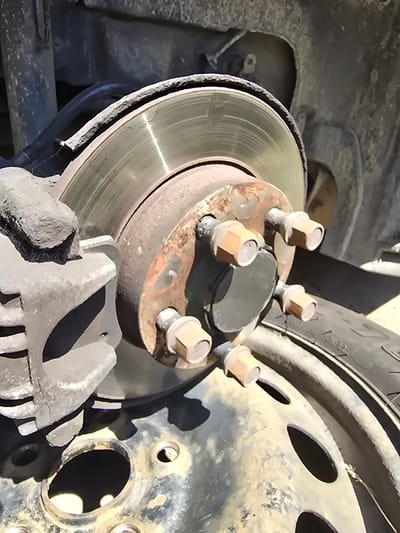This is a close-up, angled shot of a car's brake assembly, likely taken outdoors. The scene focuses on a rusty brake rotor, a gray brake caliper, and several lug nuts. The surrounding area is dusty and appears to be an unpaved surface, possibly a roadside or a parking area. The wheel of the car is partially visible, with a tire showing some tread and markings. There are no people or animals present in the image. The lighting suggests it is daytime, with harsh shadows indicating direct sunlight. The image does not contain any information that would place it in Kostelec na Hané, Czechia, or any other specific city.