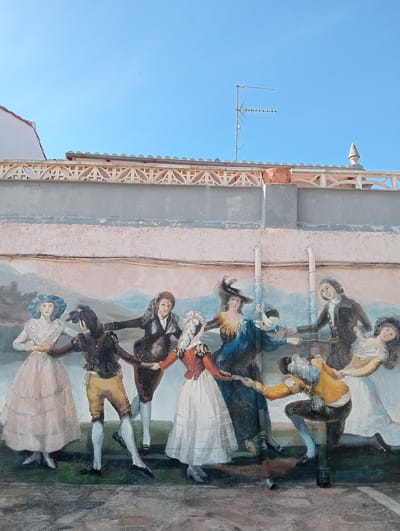 This image displays an exterior wall mural in Piedrahíta, Spain, beneath a clear blue sky. The mural depicts nine figures, both male and female, in 18th or 19th-century period attire, engaged in a circular dance while holding hands. Their expressions suggest engagement or joy. The painted background features distant rolling hills and a green foreground. Two white PVC drainpipes run vertically down the wall, partially overlaying the mural; one figure on the lower right incorporates a pipe into their posture, appearing to kneel with an arm wrapped around it. Above the mural, the wall transitions to a decorative concrete railing with geometric cutouts, topped by a tiled roof, a television antenna, and a finial. Horizontal electrical cables are visible across the wall above the artwork. The ground level comprises textured, weathered paving.