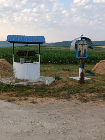 The image shows a well with a blue metal roof and a bucket hanging from a rope on the left side. To the right of the well, there is a wooden cross with a depiction of Jesus Christ and two figures, possibly Mary and John. The cross is painted blue and features decorative elements. In the background, there are fields of green crops, likely sunflowers, and a few haystacks. The ground in the foreground is a dirt road. The scene is set in an outdoor, rural environment, consistent with the outskirts of Copăceni, Moldova. The lighting suggests it is either early morning or late afternoon. There are no people visible in the image.