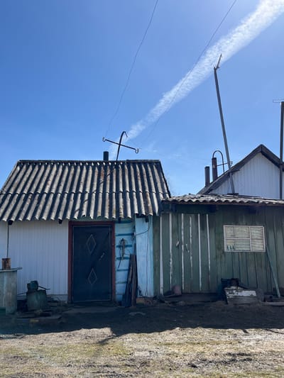 A low-angle, rotated outdoor shot depicts a multi-level building under a bright blue sky marked by a contrail.
The building features white horizontal siding at its base, transitioning to a blue-painted wall section topped by a gray, corrugated roof with an antenna. A dark door with diamond-shaped embellishments sits in the blue wall. Above, the structure shifts to green painted wood, and is topped by another section with white siding. The building is weathered, showing signs of age and disrepair.
Power lines and utility poles are visible against the sky. A patch of weedy ground borders the base of the building.
The location is Верхняя Уря, Russia, as this information was provided to me.