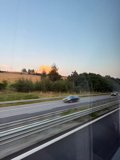 The image depicts a scenic view of a highway during dusk, as observed from a moving vehicle. The sky is a soft gradient of blue and pale orange, with a large, bright cloud formation highlighted in warm tones. The landscape features a rolling, grassy hillside dotted with scattered trees. A dense line of conifer trees forms a backdrop to the hillside. The highway itself has two lanes in each direction, separated by a median. Two cars are visible on the highway, both blurred due to motion. The foreground shows the reflective surface of what appears to be a bus or train window, with horizontal lines of the vehicle's interior structure visible, creating a sense of rapid movement. The image does not provide specific details that would allow for identification of the location within Rajana, Bulgaria.