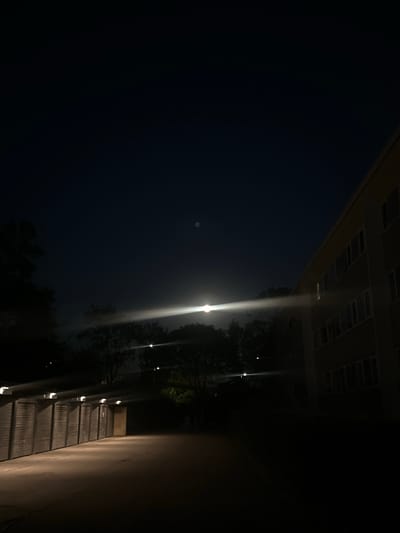 The image depicts an outdoor scene at night, likely in Roskilde, Denmark, characterized by low light conditions. On the right side of the frame, a multi-story building with a light-colored facade and numerous windows is visible. The building appears to be an apartment complex or similar residential structure. A strong beam of light, possibly from a streetlight or vehicle headlamps, cuts horizontally across the mid-section of the image, illuminating a portion of the dark sky and trees. On the left side of the frame, a row of garage doors or storage units with lights mounted above them is present. The ground in front of these units is illuminated by the overhead lights, revealing a paved surface. The overall environment suggests a residential or communal area at night, with artificial lighting providing the primary visibility. There are no people or animals visible in the image.