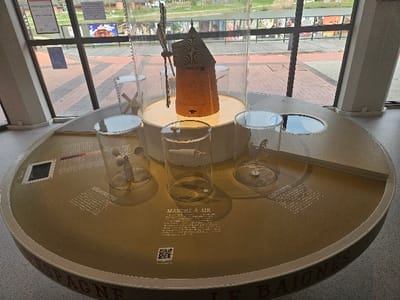 The image depicts an interior view of an exhibition space within the city of Avignonet-Lauragais, France. The primary subject is a large, circular display table, approximately three meters in diameter, with a light brown surface and white perimeter banding.
At the center of the table, a detailed model of a traditional brick-red windmill with a multi-tiered, shingled roof and four sails is prominently displayed on a raised circular platform, enclosed within a tall, clear cylindrical acrylic case.
Arranged radially around the central windmill model are three additional clear cylindrical acrylic cases, each showcasing a different weather-related instrument. The case on the left contains a three-cup anemometer. The central case houses a white, cylindrical windsock model. The case on the right displays a weather vane featuring a rooster silhouette atop an arrow. French descriptive text is engraved or printed on the table surface adjacent to each exhibit, identifying them as "ANÉMOMÈTRE," "MANCHE À AIR," and "GIROUETTE," respectively. A QR code is also visible on the table near the "MANCHE À AIR" exhibit. The outer edge of the table's banding features large red capital letters, partially visible as "SPAGNE" and "LE BAIONES."
The display table is positioned in front of a series of large, multi-paned windows with dark frames, allowing a view of the exterior. Outside, a paved area with red brick pavers arranged in a herringbone pattern is visible, bordered by a green embankment or grassy area. Beyond this, distant structures, possibly small wooden buildings with pitched roofs, and some trees are discernible under what appears to be natural daylight. Posters or signs are affixed to the exterior wall of a building across the paved area.
The interior floor is light-colored and speckled. No individuals are present in the scene.