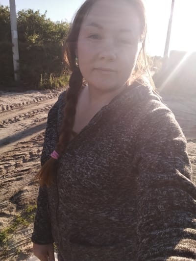 A woman is taking a selfie outdoors in what appears to be a rural area of Вазерки, Russia. She has long brown hair braided down her left side, secured with a pink elastic. She is wearing a dark, marled knit sweater or cardigan. The background shows a dirt road with tire tracks and some sparse green vegetation. Trees and a utility pole are visible to the left, with the sun shining brightly from the right, creating lens flare and washing out parts of the scene. The lighting suggests it is late afternoon or early evening. The woman's expression is neutral. There is no visible text in the image.